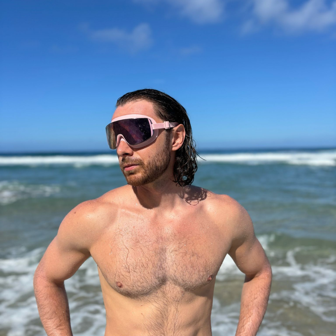 Man wearing ÆRA swimming goggles on a beach, with ocean and sky in the background, featuring anti-fog lenses and UV protection for adults