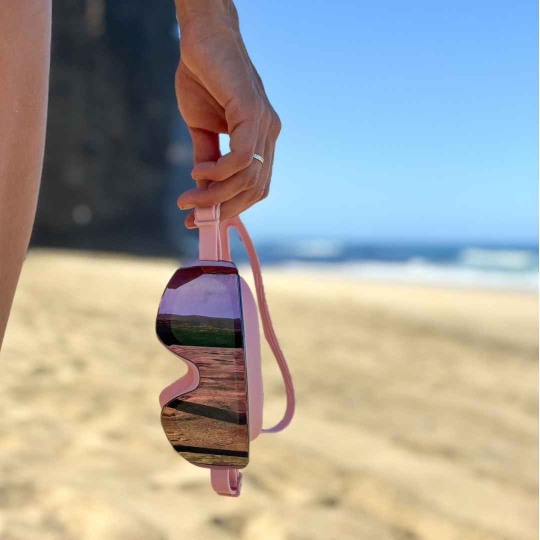Sunglasses with a pink frame held by a hand on a beach.