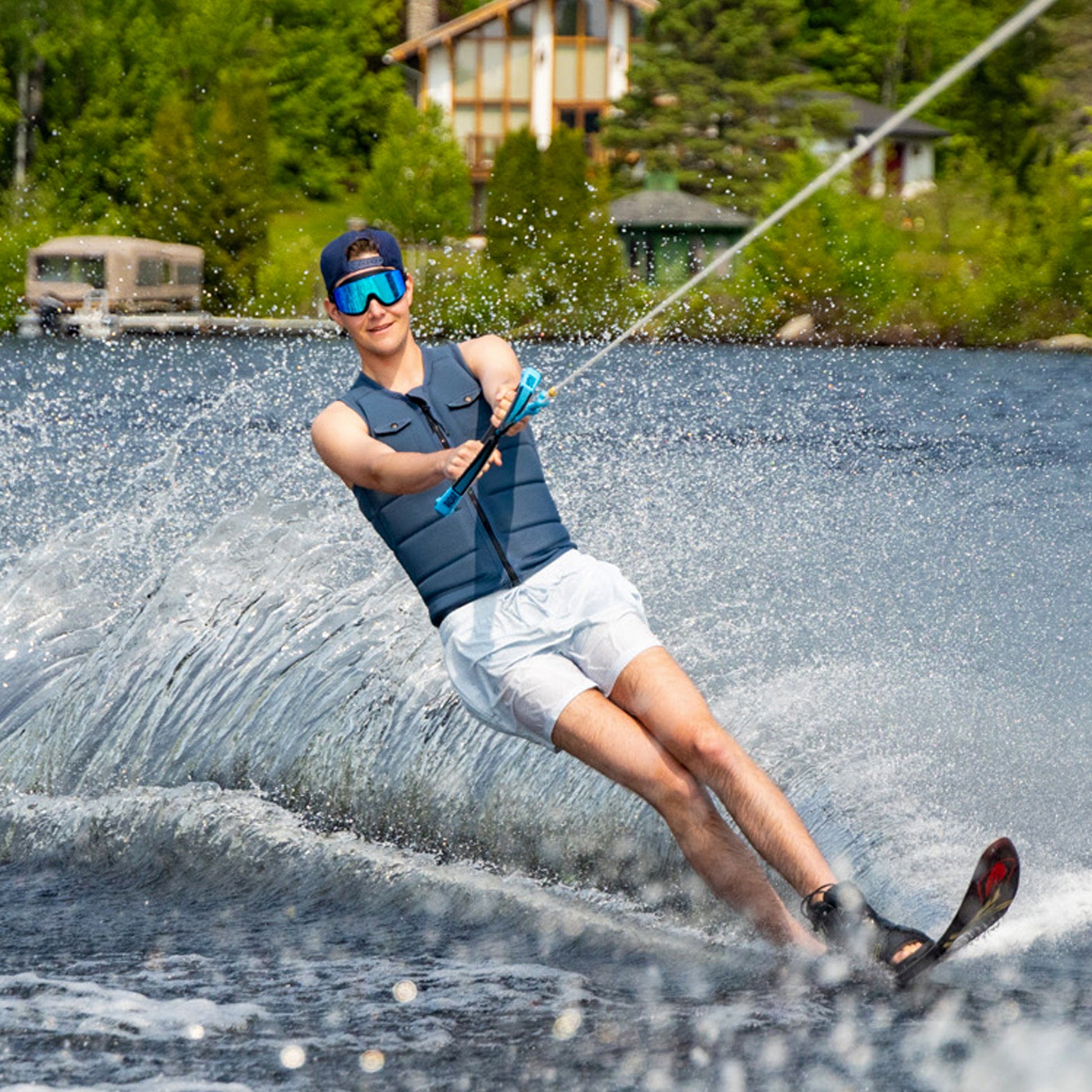 Person water skiing on a lake wearing ÆRA Blue Steel swimming goggles, with house and trees in the background, featuring anti-fog lenses and UV protection for adults