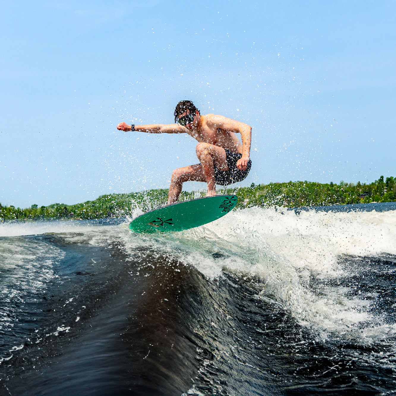Surfer riding a wave under a clear blue sky with lush greenery in the background, wearing Black Mirror AERA swim goggles for enhanced water clarity.