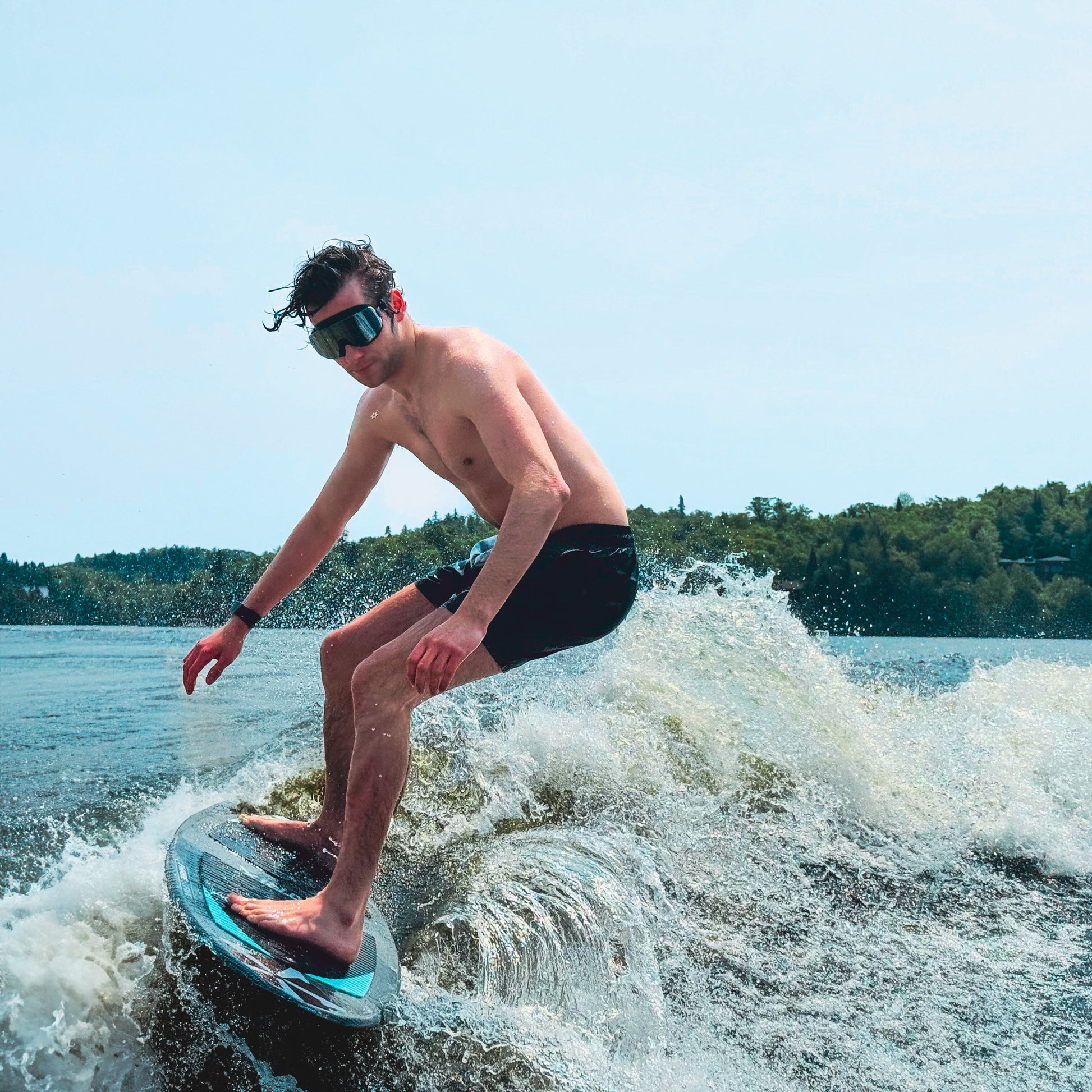 Person wakeboarding on a lake with clear sky and trees in the background, wearing Black Mirror AERA swim goggles for glare-free vision.