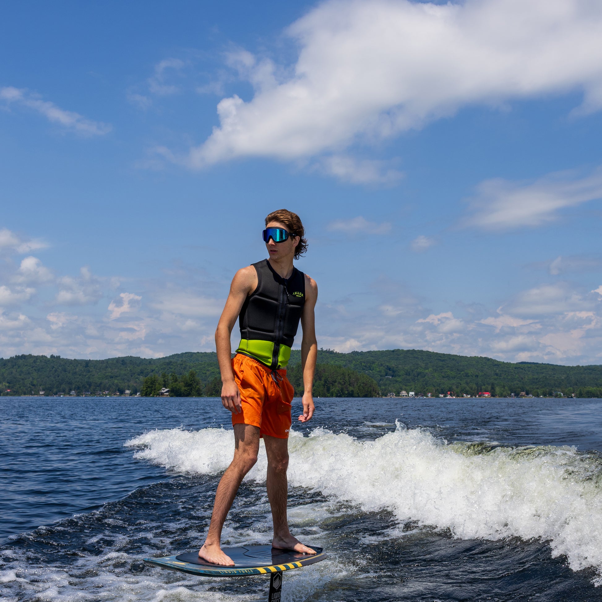 Person on a stand-up paddleboard on a lake wearing ÆRA Blue Steel swimming goggles, with blue sky and green hills in the background, featuring anti-fog lenses and UV protection for adults