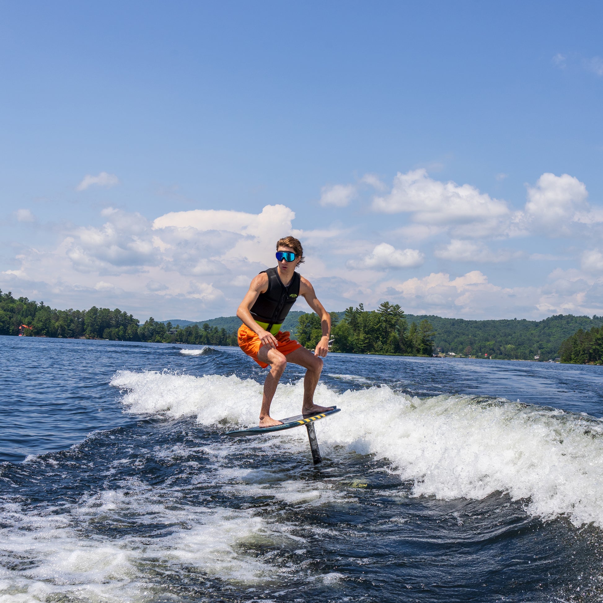 Person wakeboarding on a lake wearing ÆRA Blue Steel polarized swimming goggles, with clear blue sky and green trees in the background, featuring anti-fog lenses and UV protection for adults