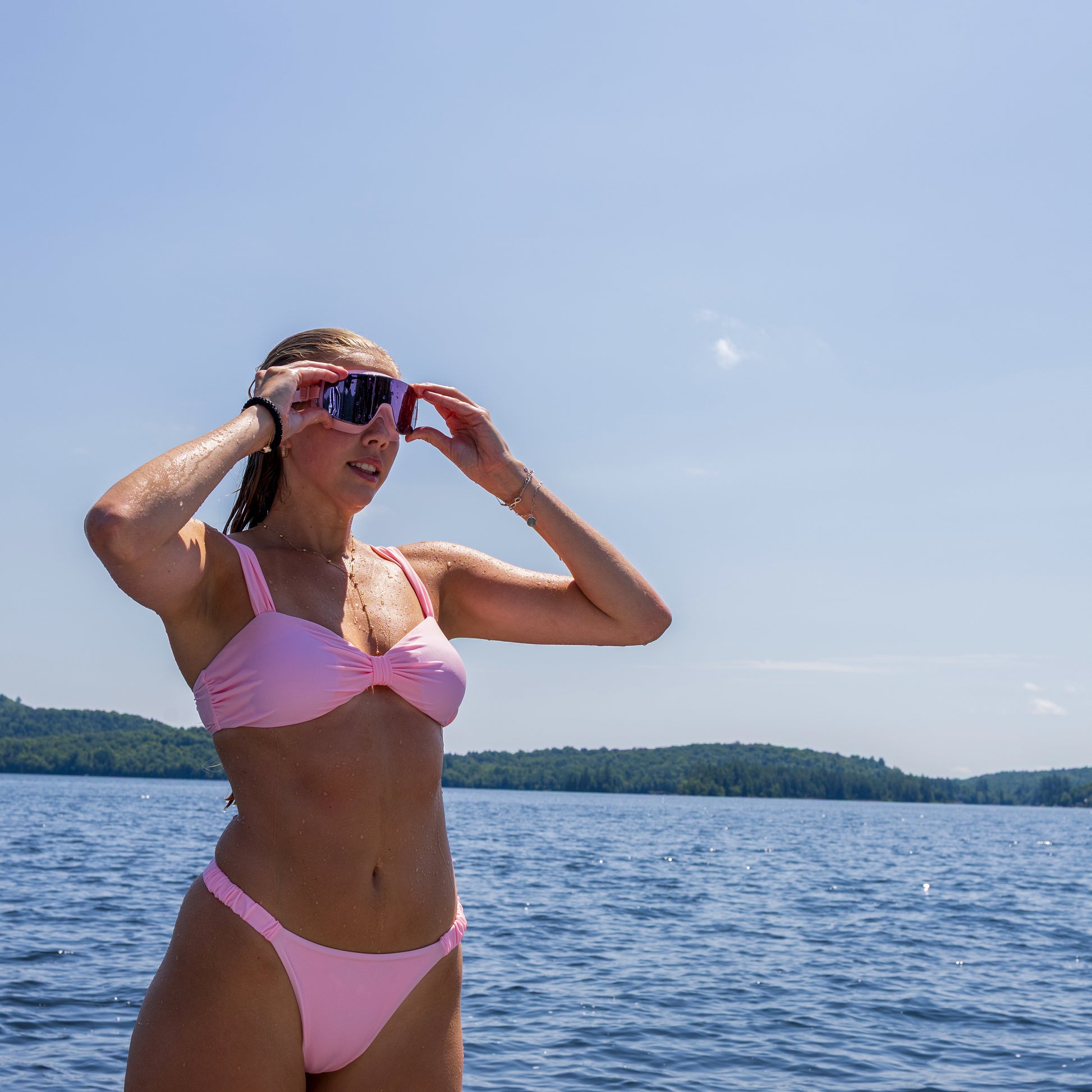 Woman in a pink bikini adjusting ÆRA Blushwave swimming goggles by a body of water, featuring anti-fog lenses and UV protection for adults