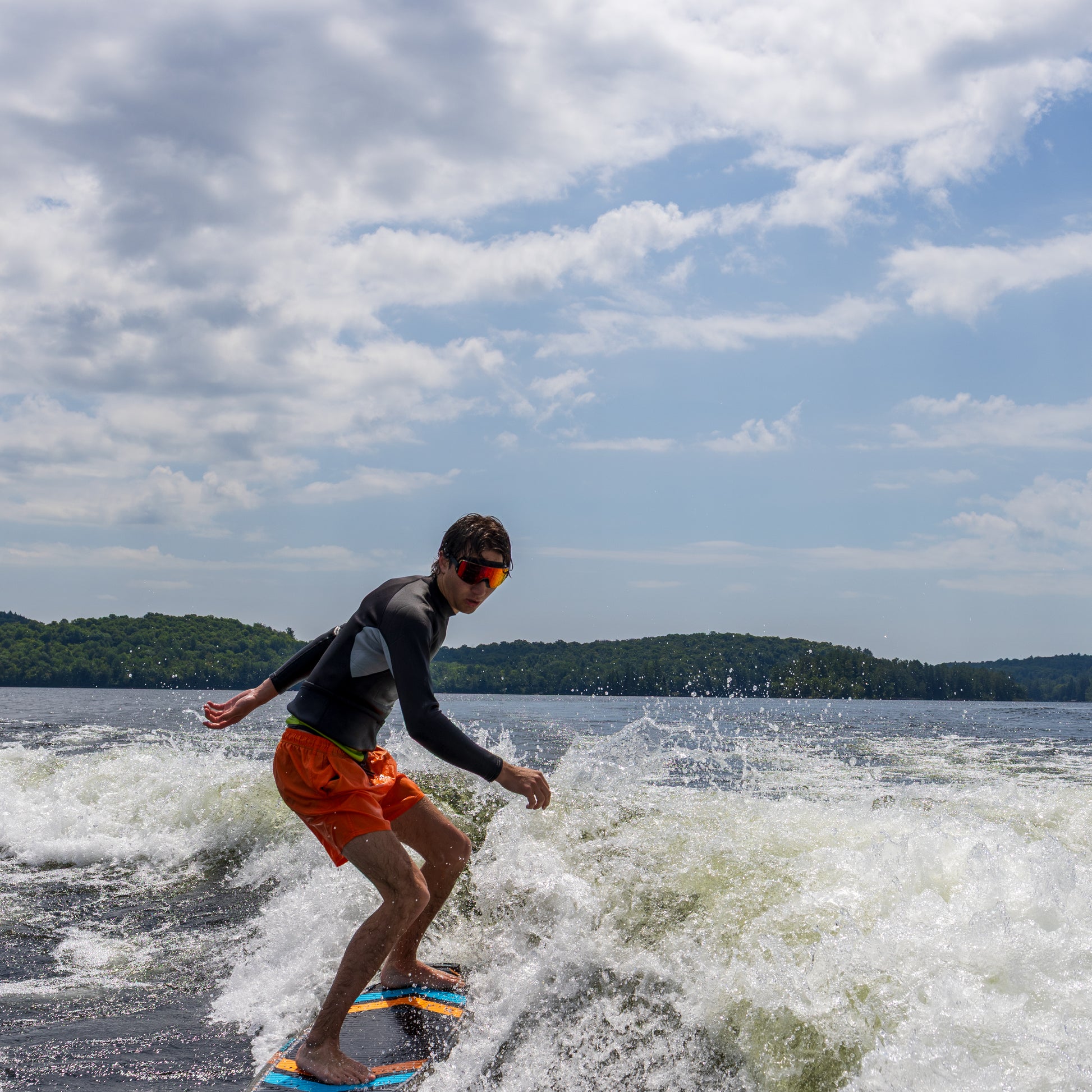 Person wakeboarding on a lake under a cloudy sky, wearing ÆRA Fire swimming goggles, featuring anti-fog lenses and UV protection for adults