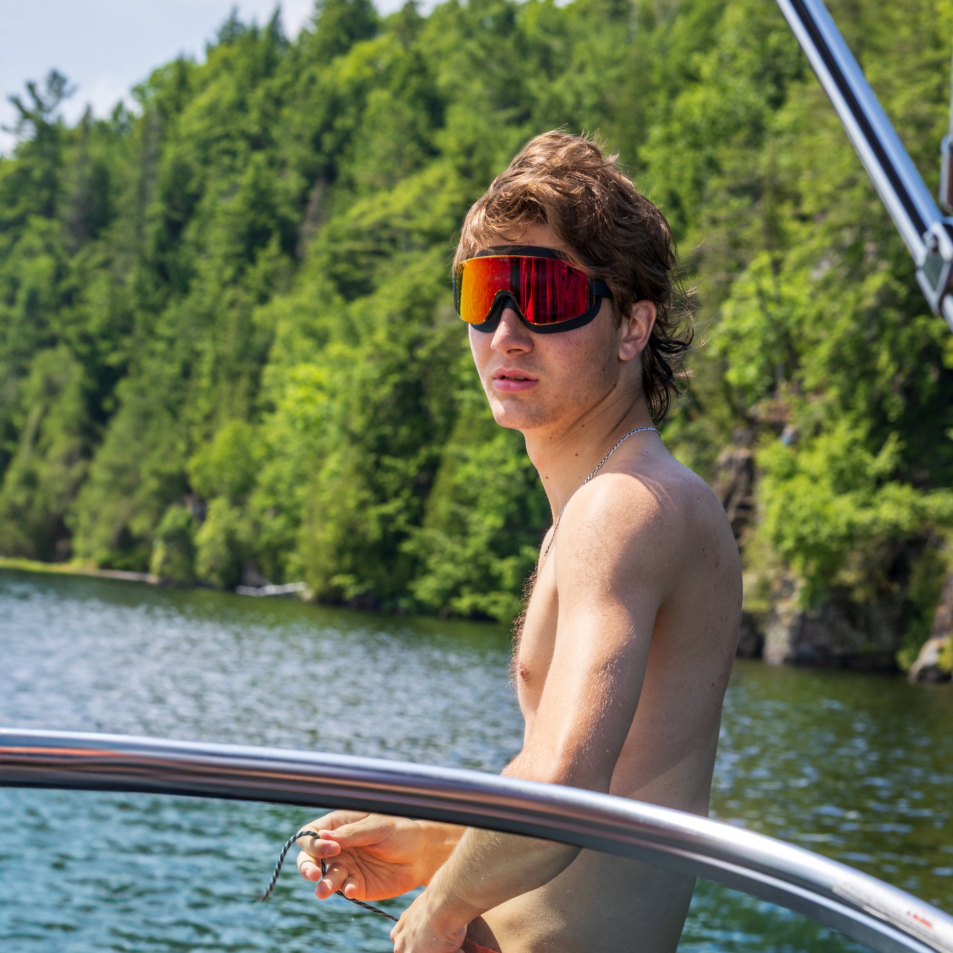Person wearing ÆRA Fire swimming goggles in red on a boat, with a forested shoreline in the background, featuring anti-fog lenses and UV protection for adults