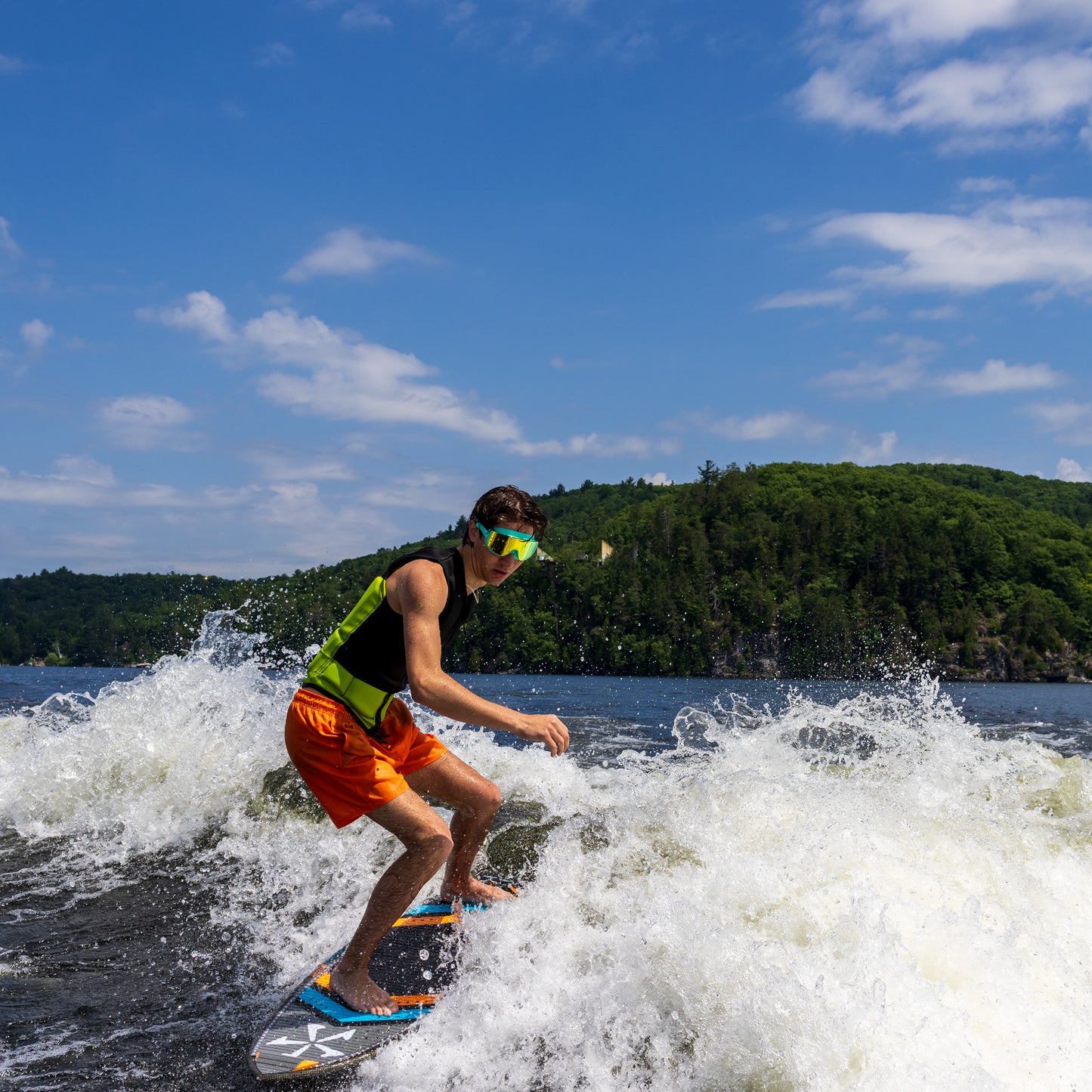 Person water skiing on a lake wearing ÆRA Emerald swimming goggles, with clear blue sky and green hills in the background, featuring anti-fog lenses and UV protection for adults