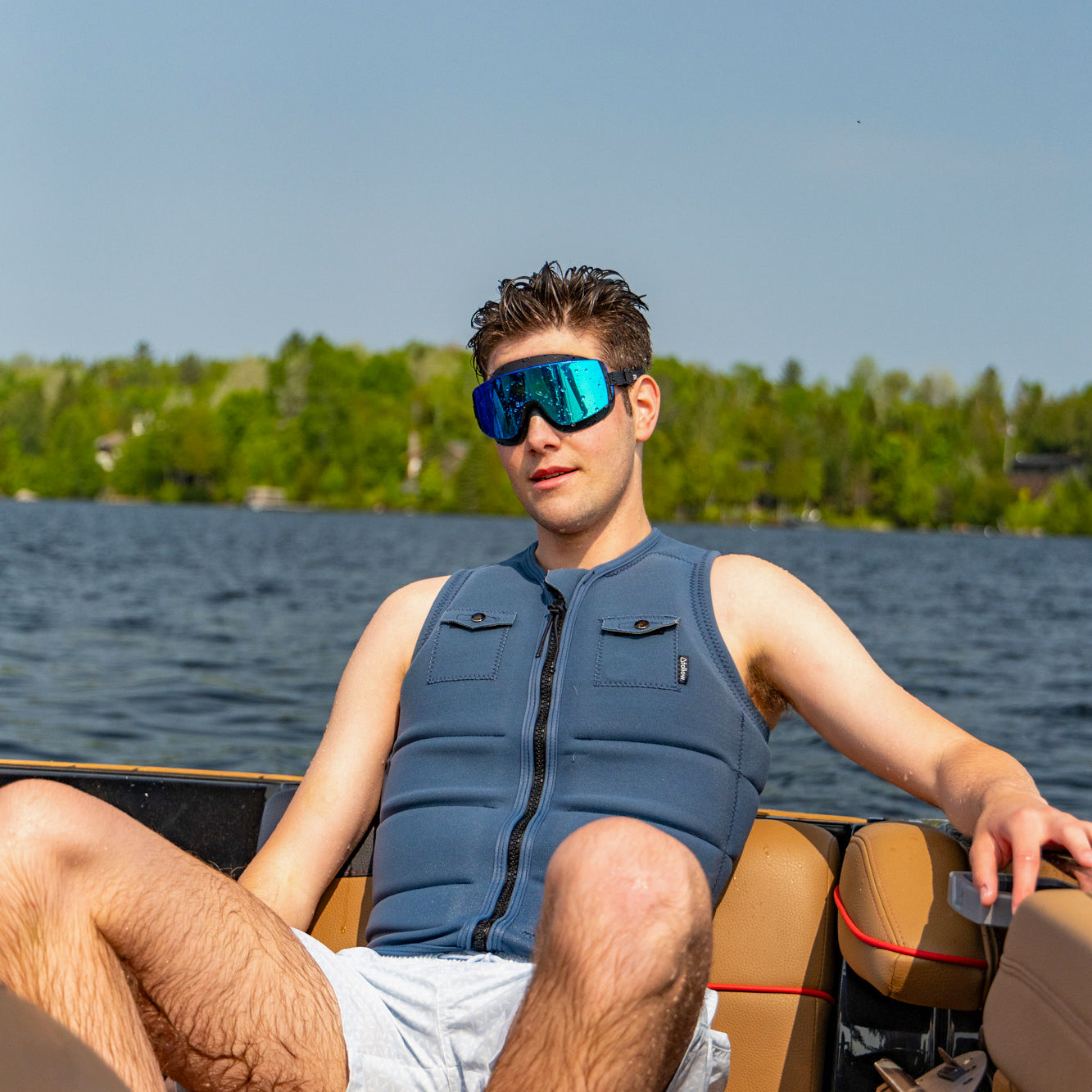 Man wearing a blue life jacket and sunglasses on a boat with a lake and trees in the background