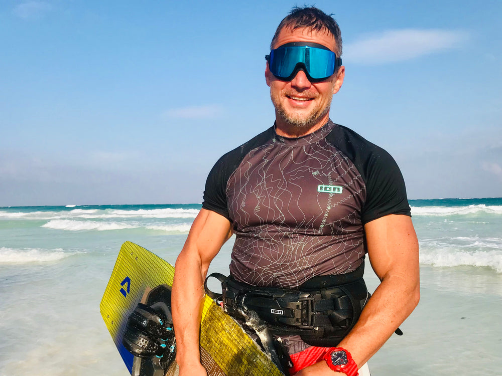 Man on a beach holding a watercraft, wearing a wetsuit and goggles.