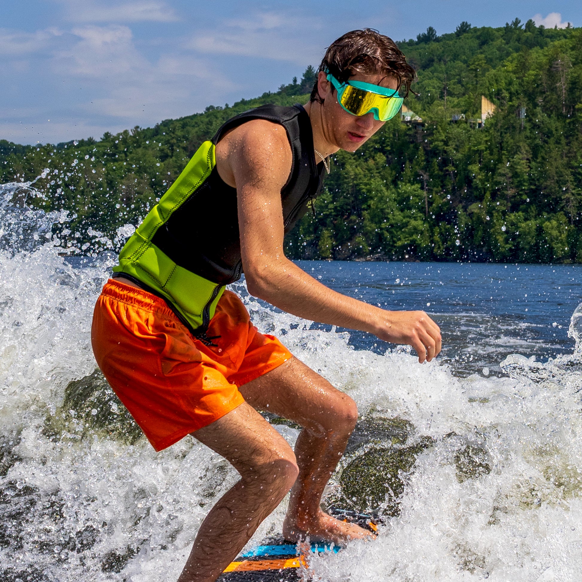 Person water skiing on a lake wearing ÆRA Emerald swimming goggles, with clear blue sky and green hills in the background, featuring anti-fog lenses and UV protection for adults