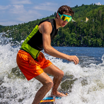 Person water skiing on a lake wearing ÆRA Emerald swimming goggles, with clear blue sky and green hills in the background, featuring anti-fog lenses and UV protection for adults