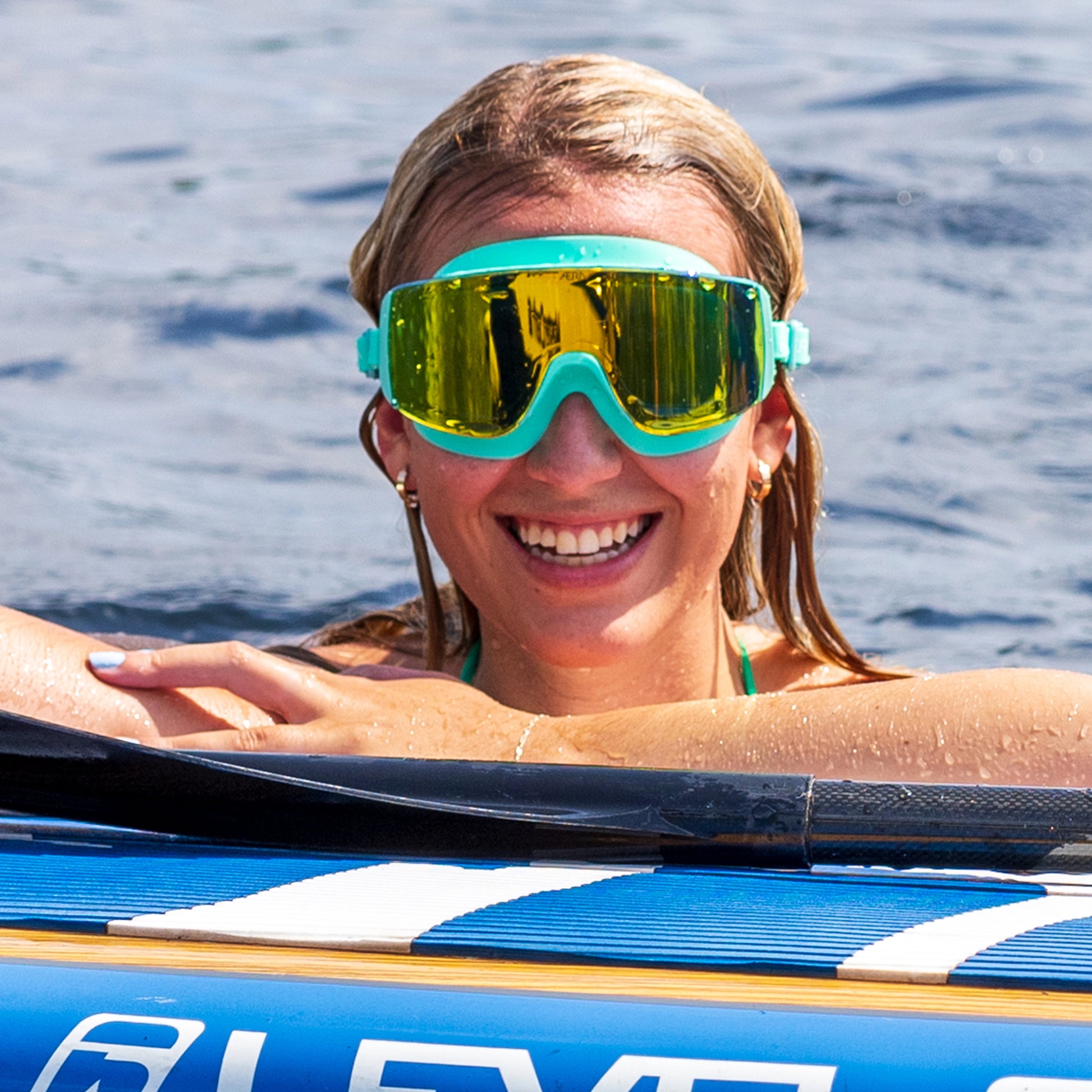Woman wearing goggles and a bikini top, sitting on a paddleboard with water in the background