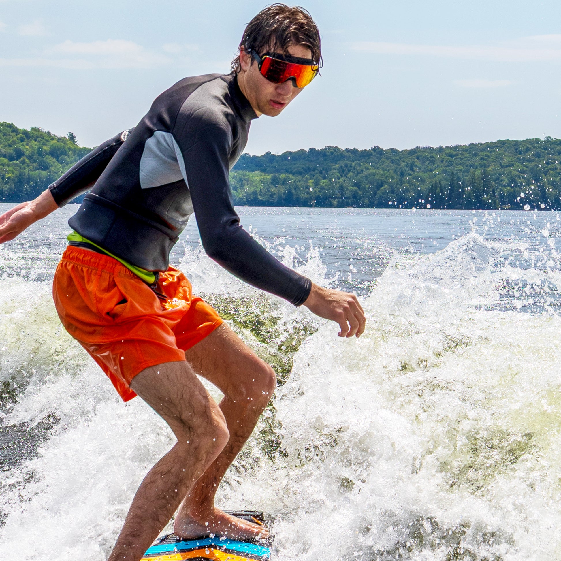 Person wakeboarding on a lake under a cloudy sky, wearing ÆRA Fire swimming goggles, featuring anti-fog lenses and UV protection for adults