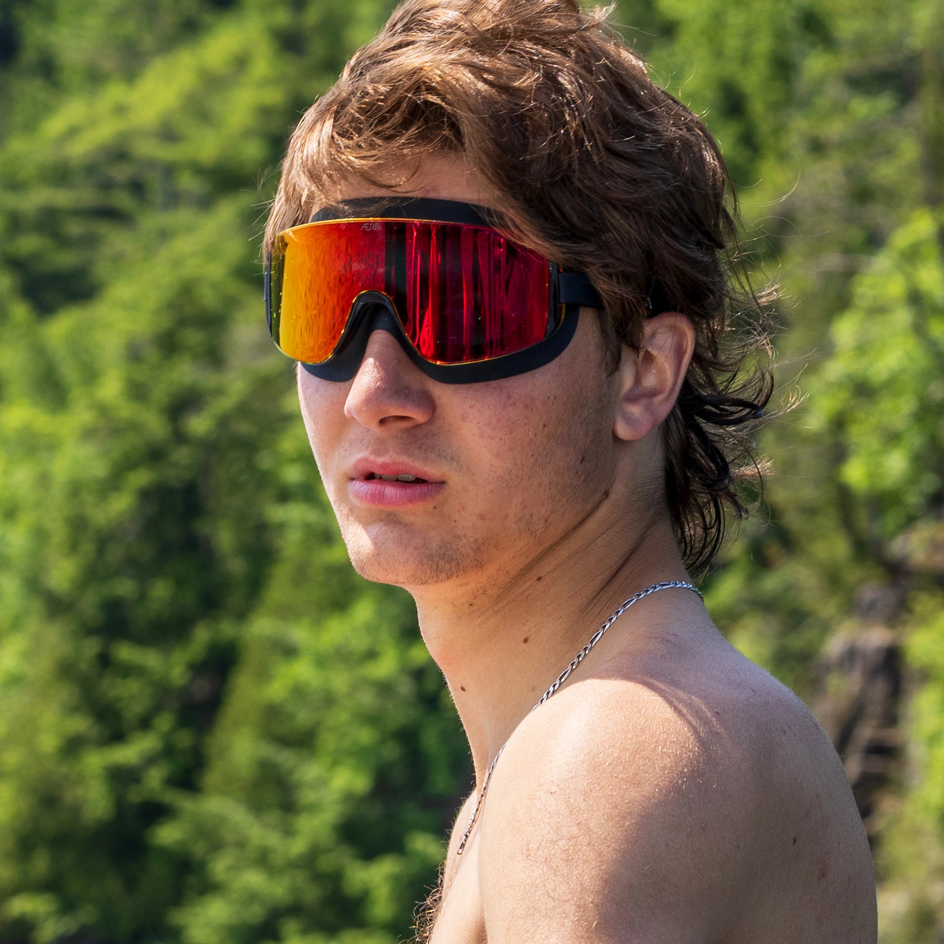 Person wearing ÆRA Fire swimming goggles in red on a boat, with a forested shoreline in the background, featuring anti-fog lenses and UV protection for adults