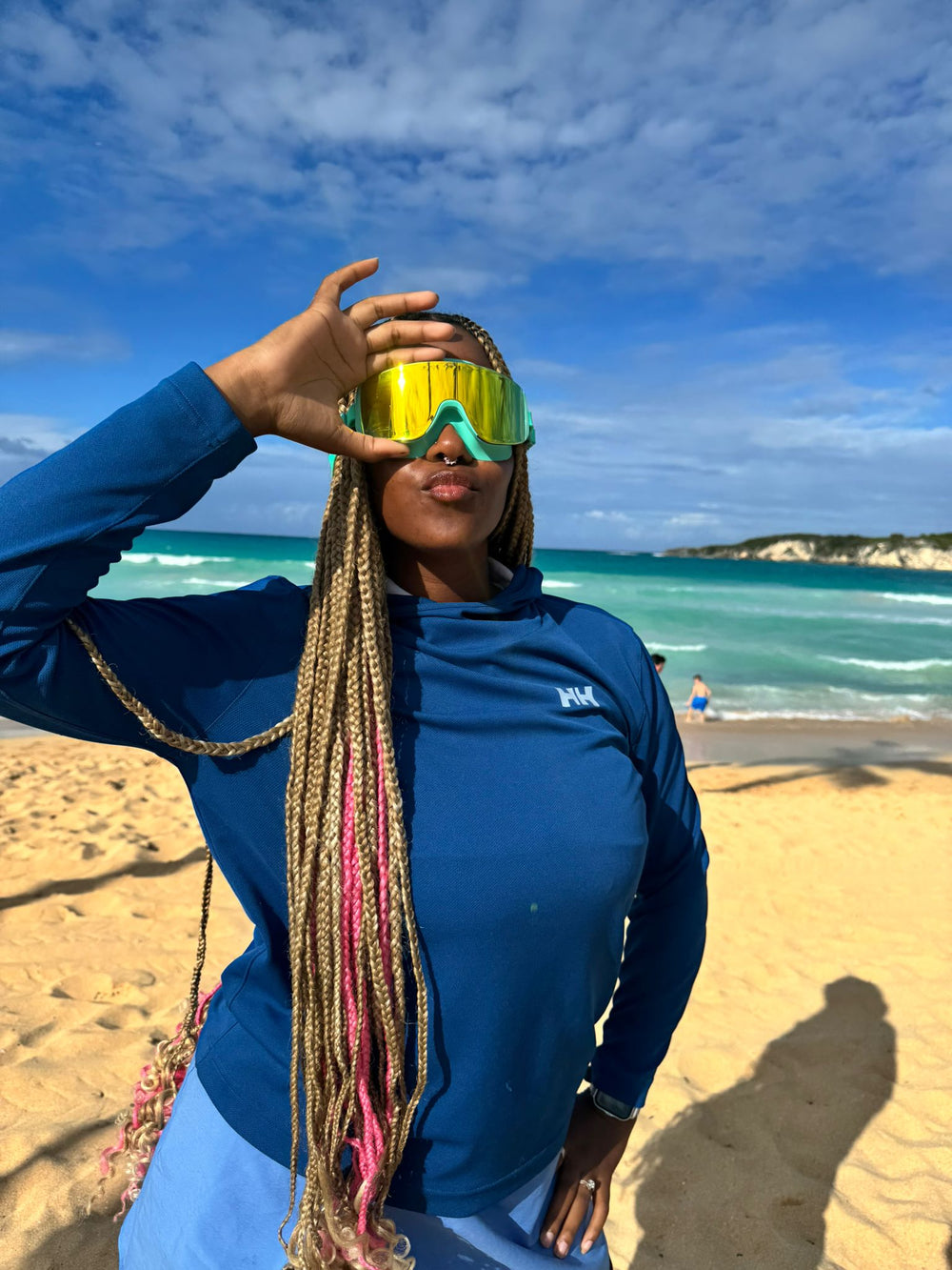 Person wearing a blue shirt and yellow goggles on a beach with ocean and sky in the background